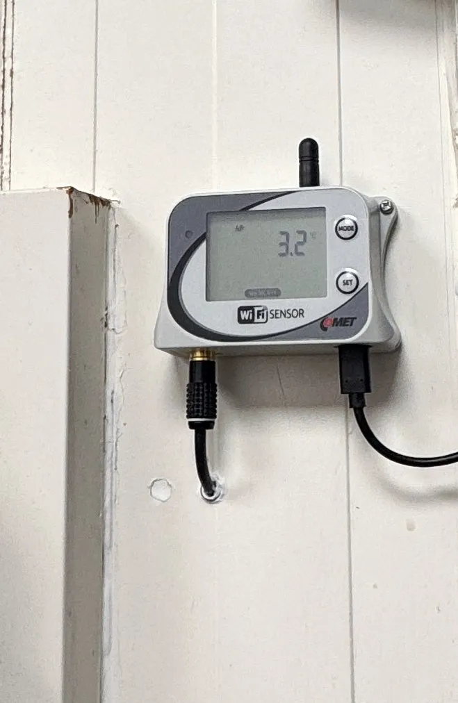 Wi-Fi datalogger Comet na stěně u vstupu do chladicího boxu, displej ukazuje 3,2 °C
